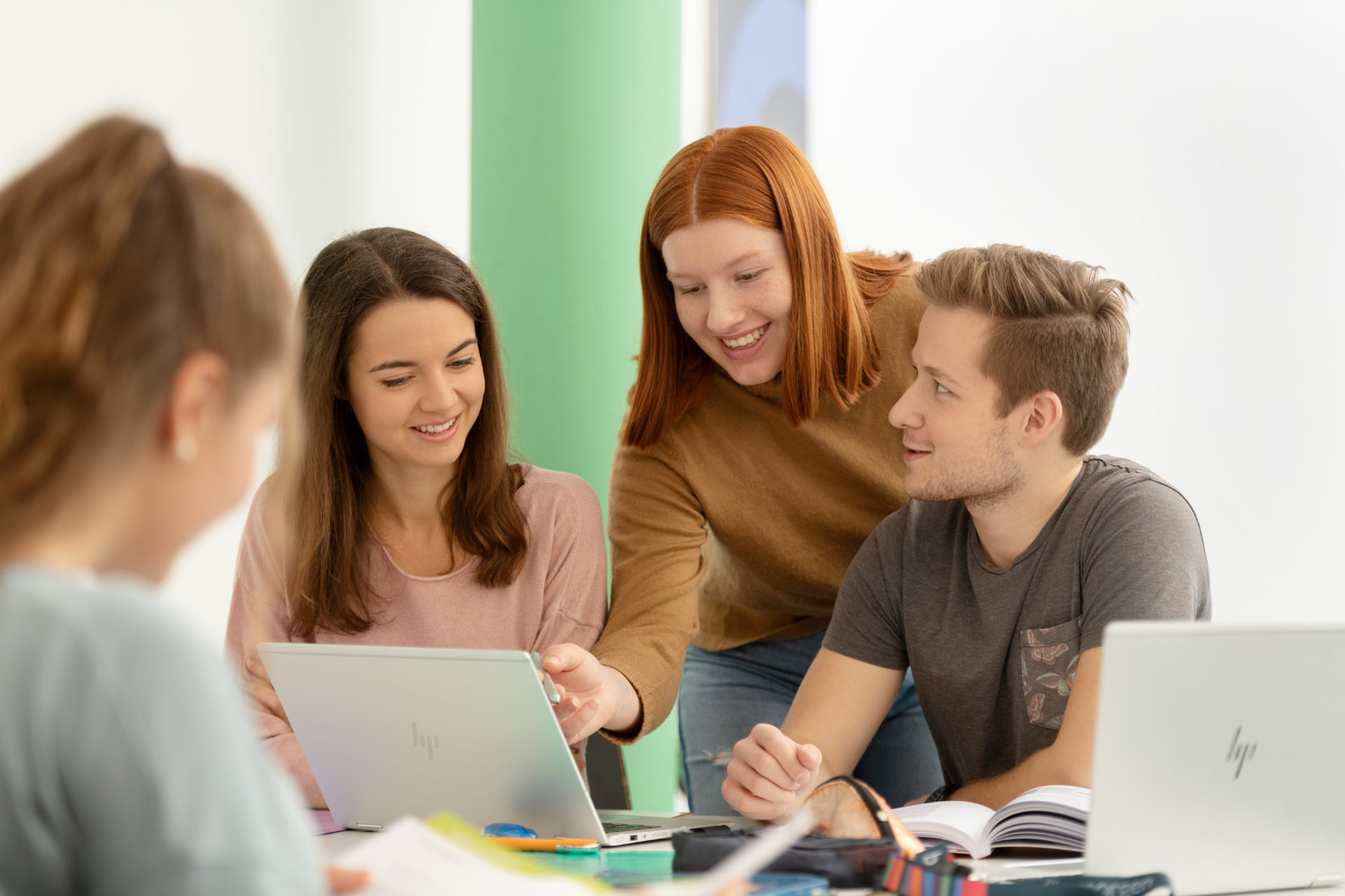 Vier Lernende um ein Pult sitzend schauen miteinander in Bücher und Laptop. Im Hintergrund eine gründe Säule und Bilder.
