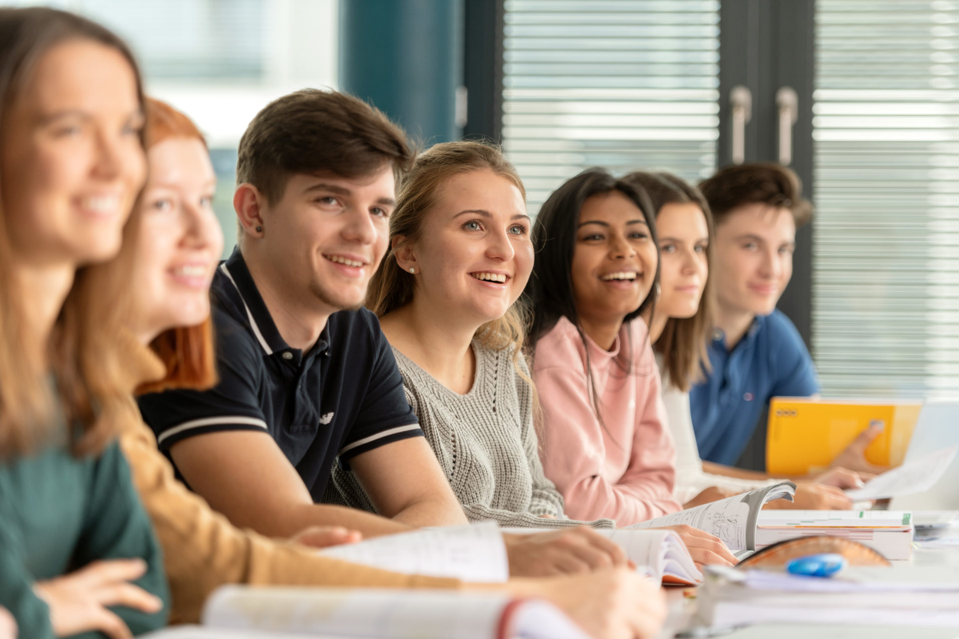 Sieben Lernende im Klassnzimmer, am Pult sitzen und nach vorne zum Lehrer schauen..