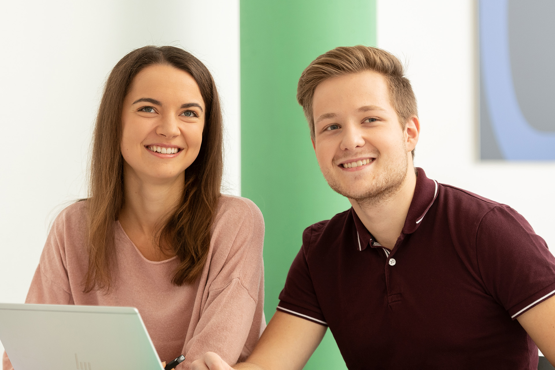Zwei Lernende vor einer grünen Säule die am Pult mit Laptop sitzen