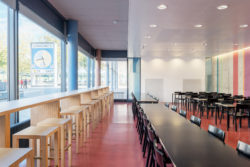 Cafeteria. Linke Seite Raumhohe Fenster mit Blick auf YB-Uhr. Davor dem Fenster entlang langer Holztisch mit Barhocker. In der Mitte schwarze Tische und Stühle vertikal hintereinander. Rechts vor Betonwand mit farbigen Streifen schwarze Tische und Stühle hintereinander.