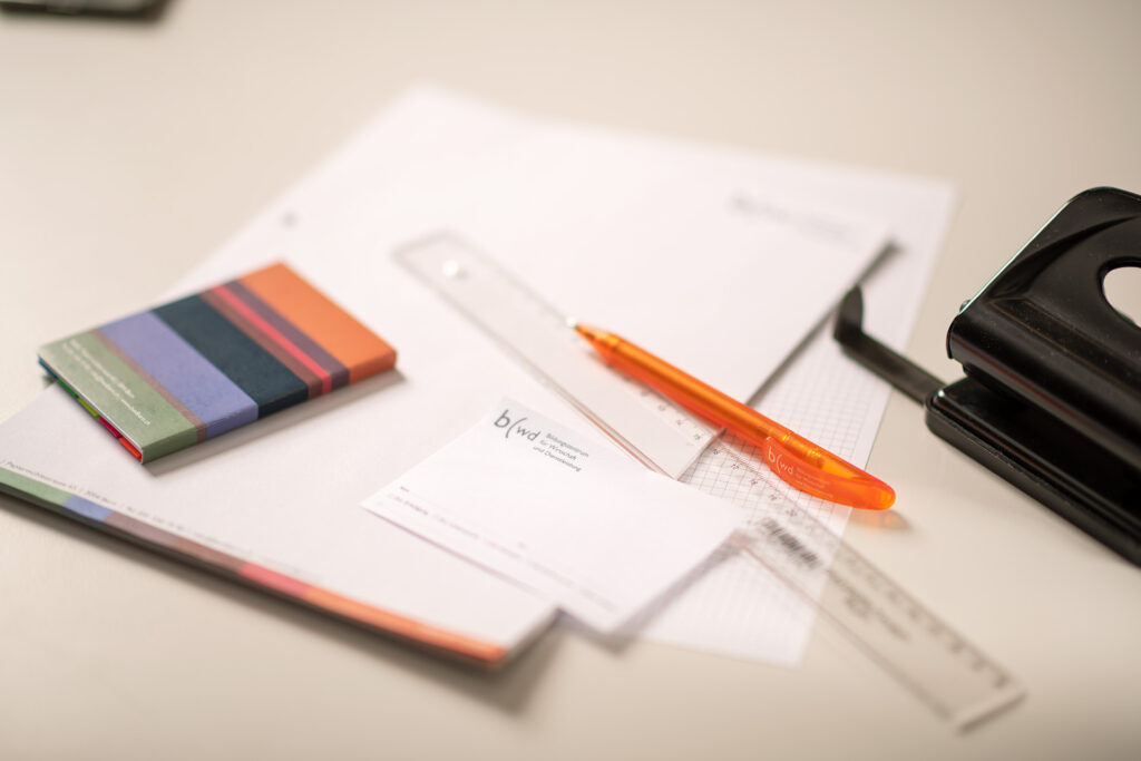 Nahaufnahme von Schreibblock, Post it-Zettel, orangem Kugelschreiber, die durcheinander auf dem Pult liegen.