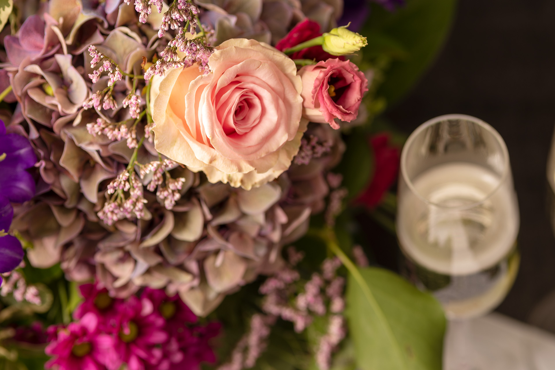 Linker Rand Blumenstrauss mit rosa, violetten und pinkfarbigen Blüten. Rechts ein Champagnerglas in der Unschärfe.